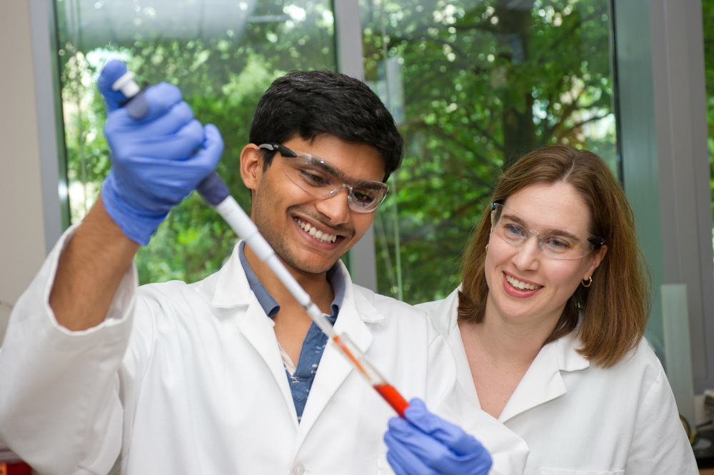 Graduate research assistant using a pipette to transfer a liquid substance into a test tube while a professor observes